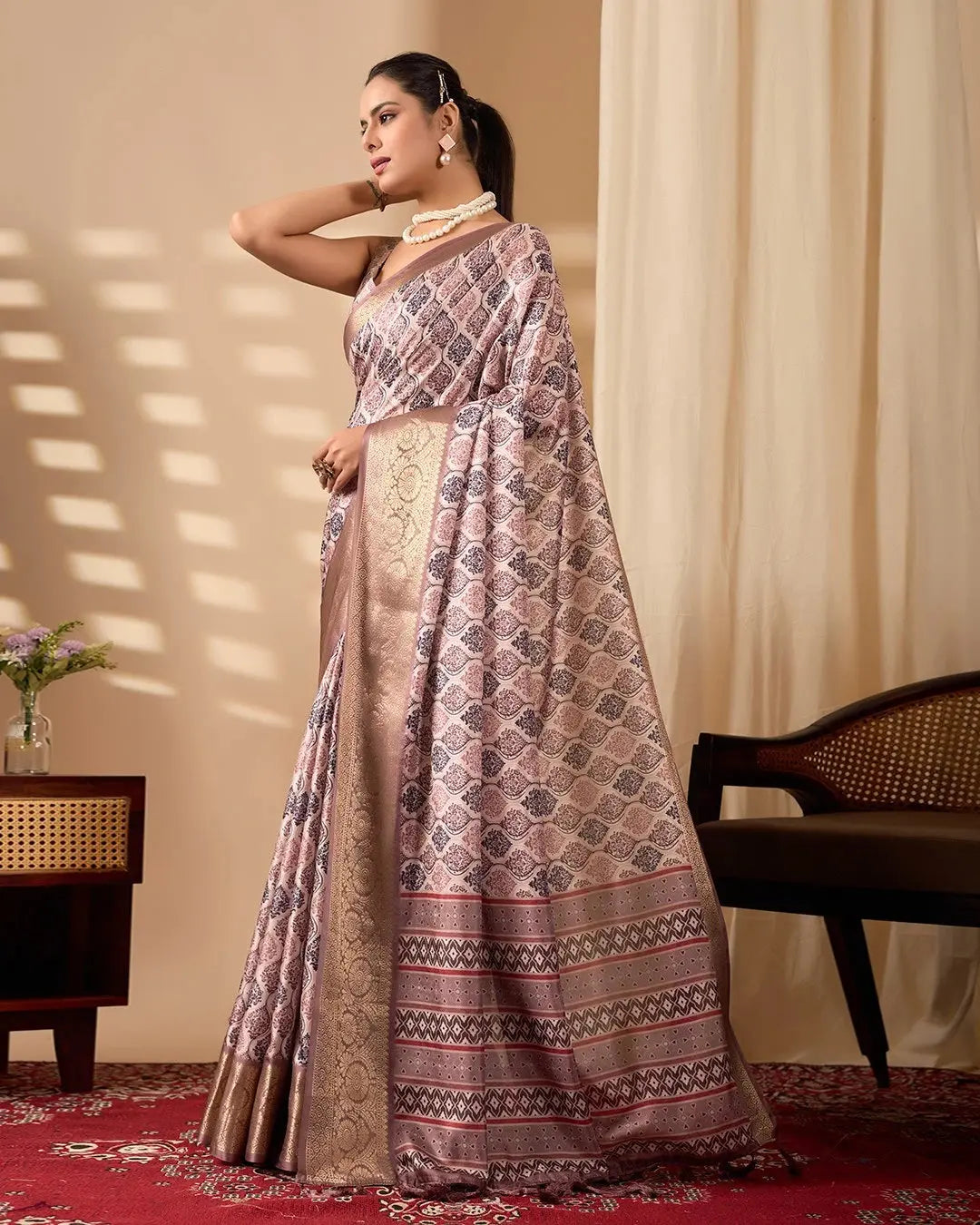 Woman wearing a traditional patterned saree in a room with a chair and table.